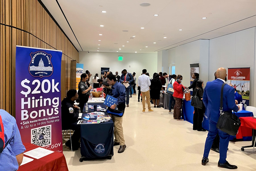 Photo of sign promoting working for the police with job seekers speaking with DC government staff behind.